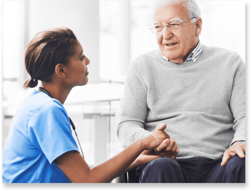 A nurse kneels in front of a patient and listens to him as she holds his hand.