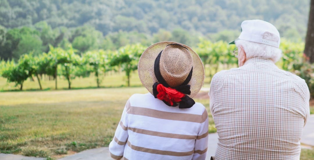 Two elderly people sit outside and look out at a park in front of them.