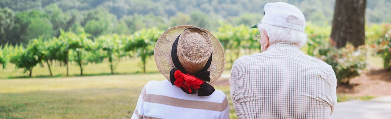 Two elderly people sit outside and look out at a park in front of them.