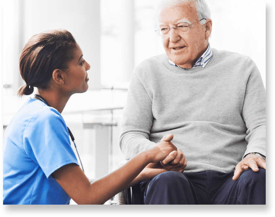 A nurse kneels in front of a patient and listens to him as she holds his hand.