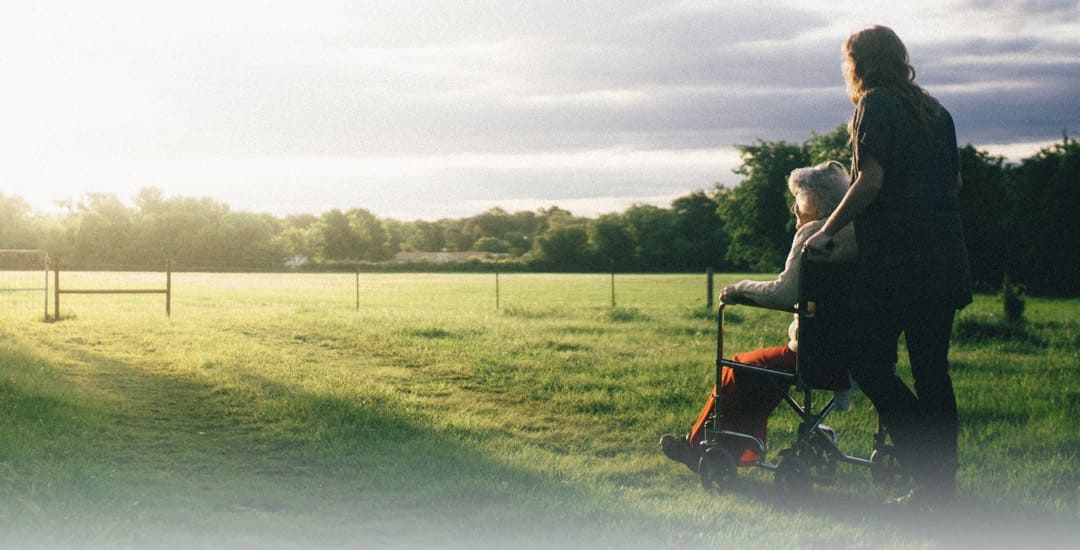 A caregiver guides a resident in a wheelchair outside in a park.