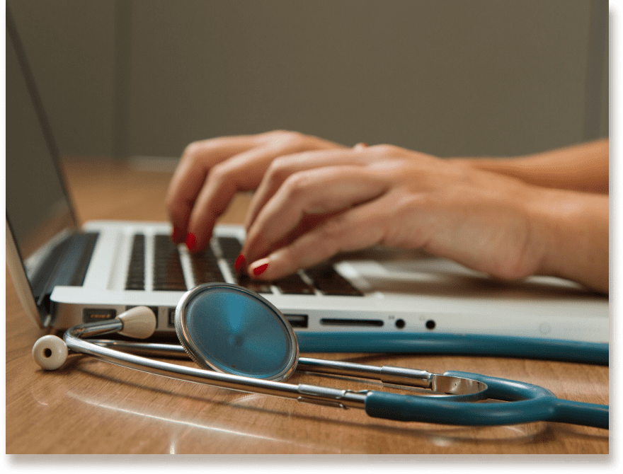 A caregiver types on a laptop with a stethoscope on the table.